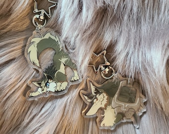 May include: Two clear acrylic keychains featuring illustrated wolf-like creatures with green and cream fur. Each keychain has a silver-toned star-shaped clasp. One charm shows a wolf in a crouching pose, while the other depicts a wolf head profile.