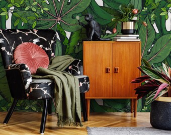 May include: A black velvet armchair with a floral pattern and a pink throw blanket sits in front of a wall with a tropical leaf wallpaper. A wooden cabinet with two doors is to the right of the chair. A small potted plant is on the floor next to the cabinet.