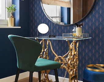 May include: A gold metal vanity table with a glass top and ornate legs. A green velvet chair sits in front of the table. The table is set against a blue wall with a geometric pattern.