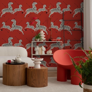 May include: A living room interior with a red wall featuring a repeating pattern of black and white zebras. The room features a white couch, two wooden end tables, a red chair, a plant, and a white shelf with books and decorative objects.