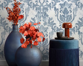 May include: Two dark blue ceramic vases with a floral arrangement of red and orange leaves. The vases are sitting on a light blue patterned background. A dark blue velvet ottoman with a fringe trim is in the foreground.