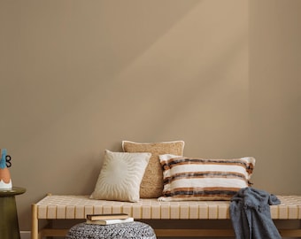 May include: A neutral-toned interior scene featuring a woven bench with three decorative pillows in beige, cream, and brown. A dark blue blanket is draped over the bench. A round, patterned ottoman and a small side table complete the cozy setting.