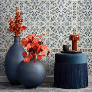 May include: Two dark blue ceramic vases with a red leafy plant in front of a white and blue patterned wallpaper. A blue velvet ottoman with a fringe trim sits in front of the vases.