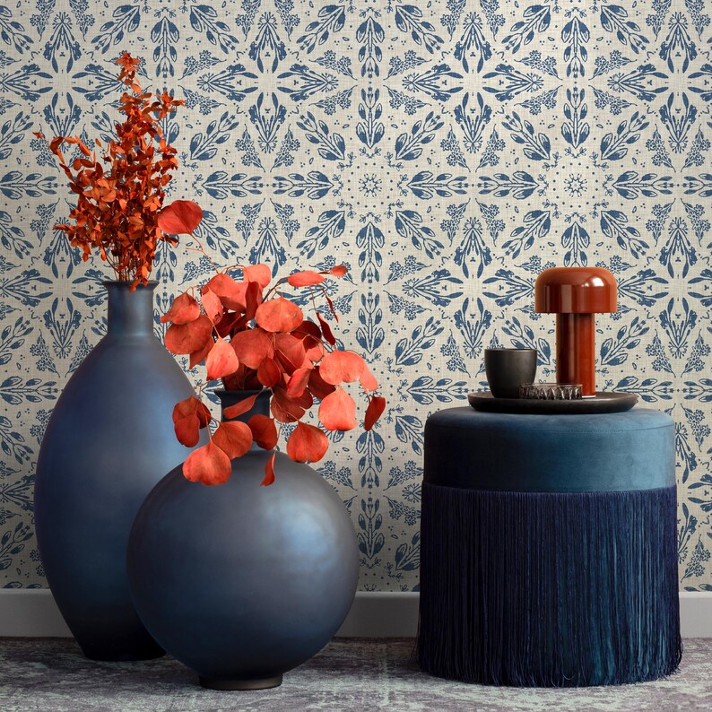 May include: Two dark blue ceramic vases with a red leafy plant in front of a white and blue patterned wallpaper. A blue velvet ottoman with a fringe trim sits in front of the vases.
