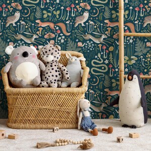 Puede incluir: Una cesta de almacenamiento de mimbre llena de animales de peluche, incluyendo un koala, un leopardo, un conejito y un ping&uuml;ino. La cesta est&aacute; delante de un papel tapiz azul verdoso con una escena de bosque caprichosa con animales, setas y plantas.