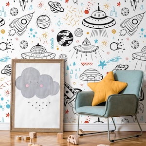 May include: A whimsical space-themed nursery decor scene. The white wall features black and white illustrations of rockets, planets, and UFOs. A framed picture of a smiling gray cloud with raindrops sits near a blue armchair with a yellow star-shaped pillow.