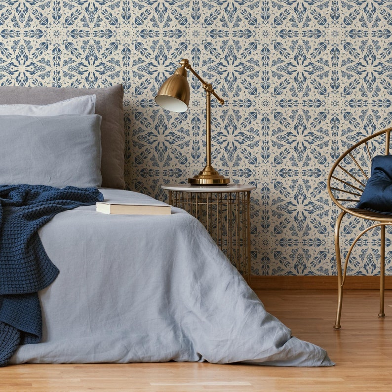 May include: A blue and cream patterned wallpaper with a geometric design. A bed with a blue blanket and pillows is in the foreground. A gold lamp and side table are next to the bed. A gold chair with a blue pillow is in the background.