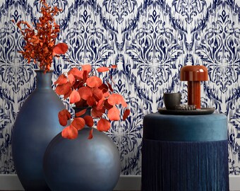 May include: Two dark blue ceramic vases with a floral arrangement of red dried branches against a white and blue patterned wallpaper. A blue velvet ottoman with a fringe trim sits in front of the vases.