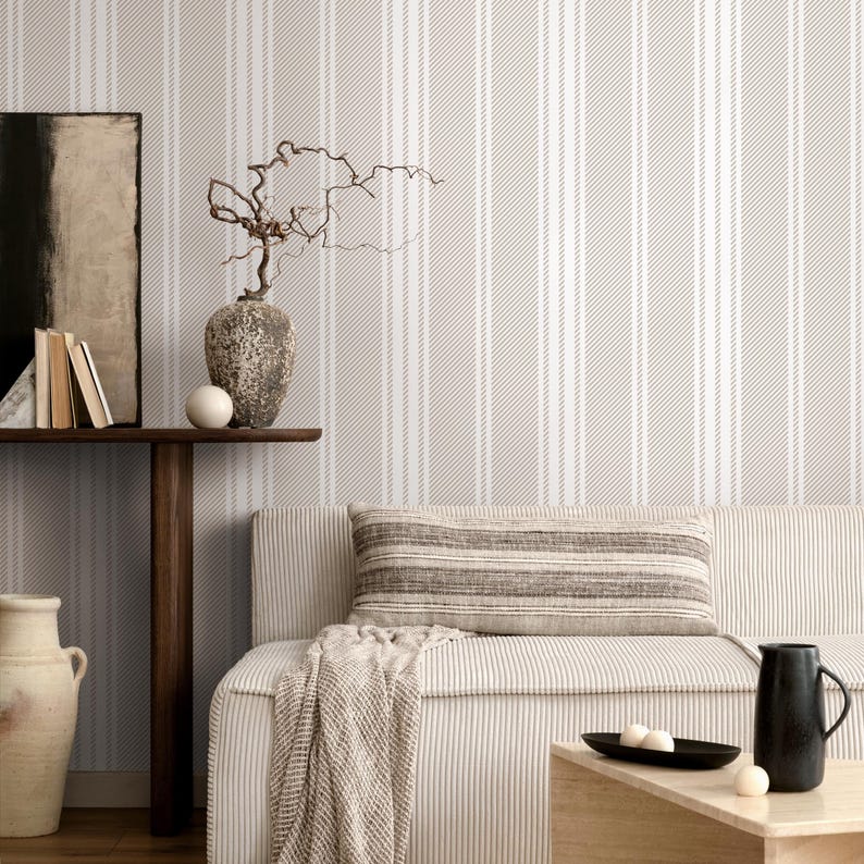 May include: A light beige striped wallpaper with a white stripe pattern. The wallpaper is in a living room with a couch, a throw blanket, and a vase with a branch.