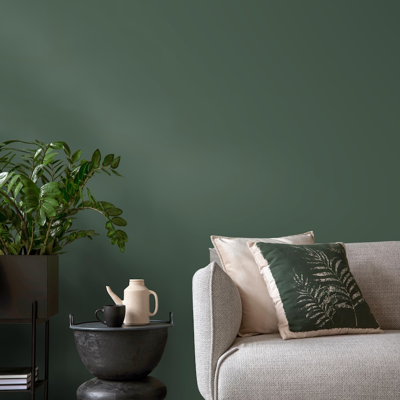 May include: A gray sofa with a white and green patterned pillow. The sofa is in front of a dark green wall. A small black side table with a ceramic teapot and a cup sits in the foreground.