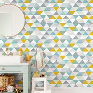 Può includere: Un bagno con una carta da parati geometrica con triangoli nei toni del giallo, grigio, verde acqua e bianco. La stanza comprende uno specchio rotondo, un lavandino bianco e un mobiletto blu chiaro. Sono visibili anche un portasciugamani dorato e uno sgabello bianco.