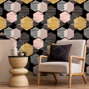 May include: A modern interior scene featuring a geometric wallpaper with a repeating pattern of black, gold, pink, and white hexagons. A beige armchair with wooden arms and a decorative pillow sits in front of the wallpaper. A small wooden side table and a white vase are also visible.