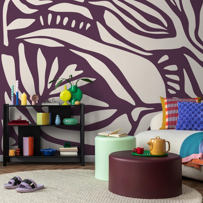 May include: A living room with a purple and white abstract patterned wallpaper, a white couch with colorful pillows, a black shelf with colorful decorative objects, and two round, colorful ottomans.