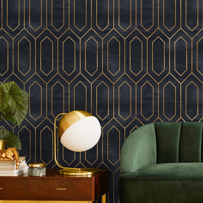 May include: A gold table lamp with a white globe shade sits on a wooden table in front of a dark blue wall with a gold geometric pattern. A dark green velvet sofa is in the background.