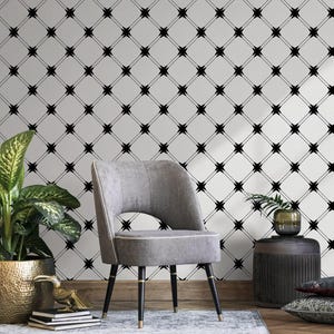 May include: A gray upholstered chair with black legs and gold accents sits in front of a wall with a black geometric pattern. A potted plant and a small side table with a plant are also in the room, creating a modern interior design.