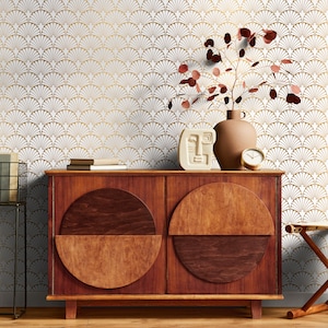 May include: A wooden dresser with two doors and two half-circle pulls. The dresser is in front of a white wall with a gold geometric pattern. A brown vase with a dried plant and a white ceramic sculpture are on top of the dresser.