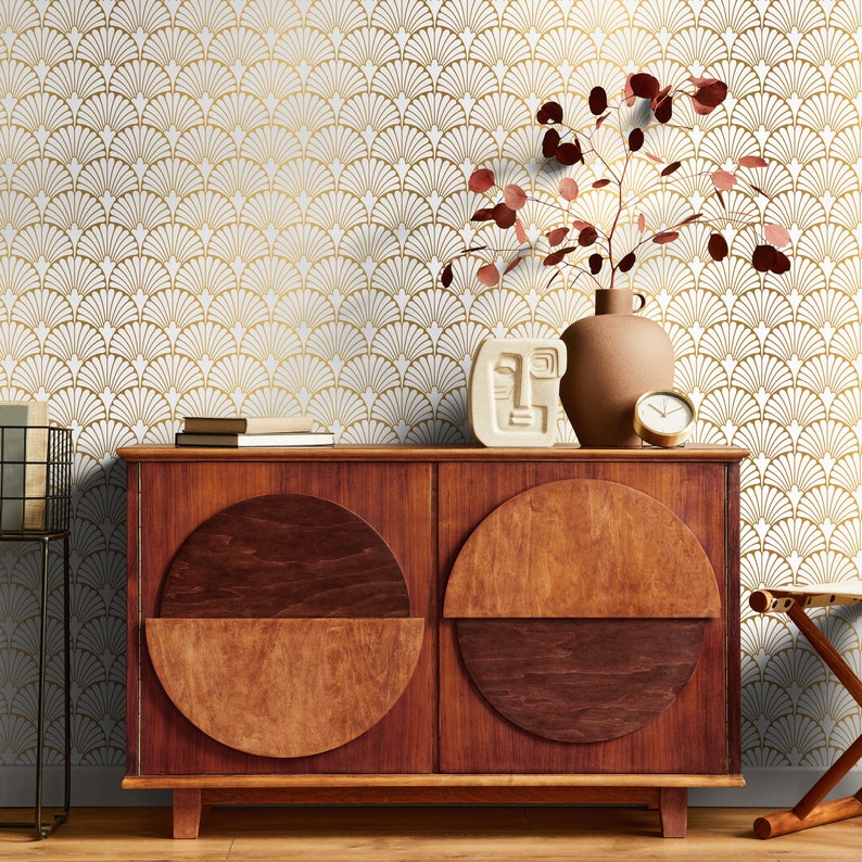 May include: A wooden dresser with two doors and two half-circle pulls. The dresser is in front of a white wall with a gold geometric pattern. A brown vase with a dried plant and a white ceramic sculpture are on top of the dresser.