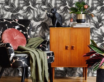 Puede incluir: Un sillón negro con estampado floral y una manta verde se encuentra frente a un papel tapiz floral blanco y negro. Un armario de madera con dos puertas se encuentra a la derecha del sillón. Una figura de mono negro se encuentra encima del armario. Una planta en maceta con hojas verdes y flores rosas se encuentra encima del armario.
