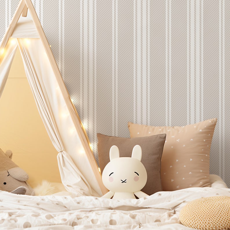 May include: A white stuffed bunny toy sits on a bed with a white and gray striped wallpaper. The bed has a white comforter and a brown pillow.