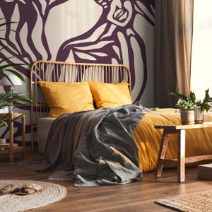 May include: A bedroom with a mustard yellow bedspread and a dark gray throw blanket. The headboard is made of woven wood and the wall behind the bed is decorated with a large abstract floral mural in shades of purple and white.