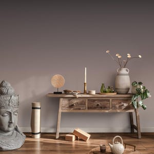 May include: A serene interior scene featuring a wooden console table adorned with decorative items. A large gray Buddha head statue is on the left. The table holds a vase with dried flowers, candles, and small glass bottles. A yoga mat and blocks are also present.