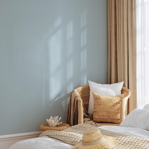 Könnte beinhalten: Eine hellblaue Wand mit einem Fensterschatten, ein Korbstuhl mit Kissen und einer Decke sowie ein Strohhut auf einem Bett. Ein kleiner gewebter Tisch hält ein dekoratives Korallenstück.