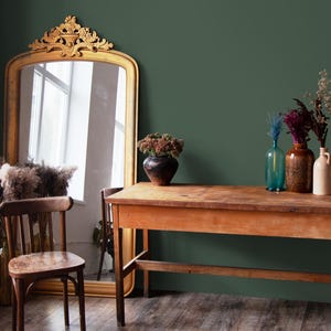 May include: A wooden table with a rustic finish sits in front of a green wall. A gold framed mirror with an ornate design reflects a window.  Vases with dried flowers and a potted plant are on the table.