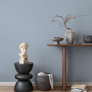 May include: A decorative interior scene featuring a white bust sculpture atop a black, stacked stone-like pedestal. A wooden console table displays a vase with branches, a small bowl, and a dark bowl. A dark pot with a folded blanket is also present.
