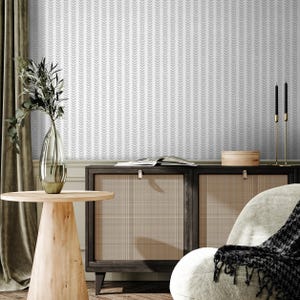 Puede incluir: Interior de una habitación con una mesa auxiliar de madera clara con un jarrón con vegetación, un mueble de madera oscura y un sillón blanco con una manta estampada en blanco y negro. El papel pintado tiene un patrón de flechas grises repetido.
