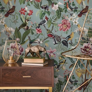 Könnte beinhalten: Eine türkisfarbene Blumentapete mit Vögeln und Blumen bildet den Hintergrund für einen Nachttisch aus dunklem Holz mit einer goldenen Drahtlampe und einer geometrischen Bärenskulptur. Rechts ist ein goldener Drahtstuhl.