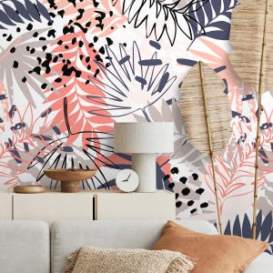 Op de afbeelding: Een kamerinterieur met een witte kast, een beige bank en een decoratieve muur met een bloemenpatroon. Het bloemmotief bevat koraal-, grijze, zwarte en marineblauwe bladvormen. Een tafellamp en een klok staan op de kast.