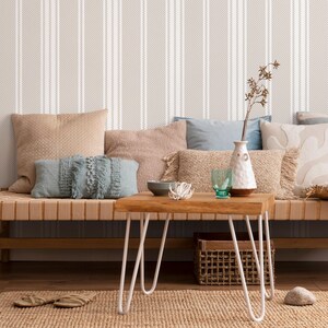 May include: A light brown couch with a variety of throw pillows, including a blue pillow with fringe, sits in front of a wall with a striped pattern. A small wooden table with white hairpin legs sits in front of the couch. A vase with a white ceramic finish and a glass of water are on the table.