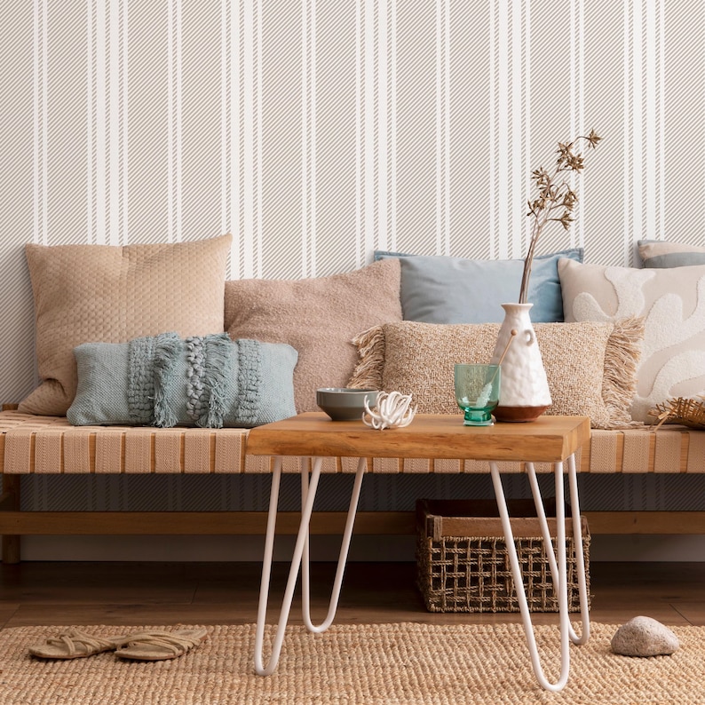 May include: A light brown couch with a variety of throw pillows, including a blue pillow with fringe, sits in front of a wall with a striped pattern. A small wooden table with white hairpin legs sits in front of the couch. A vase with a white ceramic finish and a glass of water are on the table.