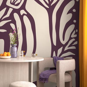 May include: A dining room with a white wall featuring a large abstract purple and white floral design. A round wooden table with two glasses and a vase of purple flowers sits in front of the wall. A grey chair with a purple throw blanket sits next to the table.