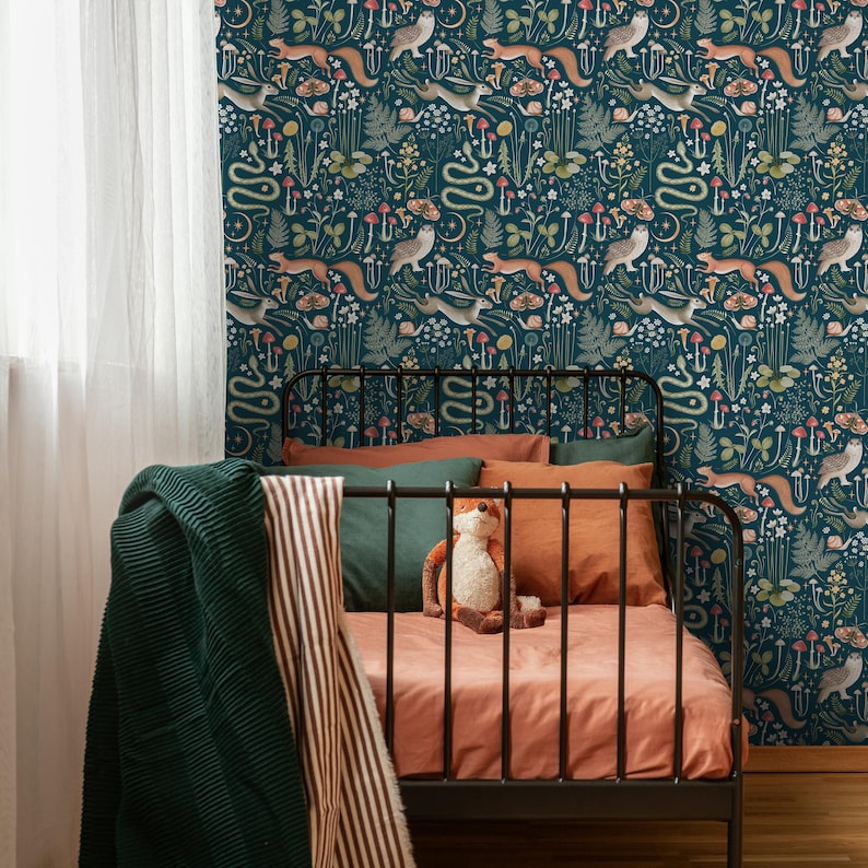 Puede incluir: Una cama infantil azul oscuro con un marco de metal negro y un papel tapiz estampado con animales del bosque, flores y setas. La cama tiene un edred&oacute;n de color melocot&oacute;n y una manta a rayas verde y blanca.