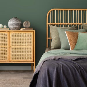 May include: A rattan headboard with a green and brown patterned throw blanket on a bed with a dark gray blanket. A wooden cabinet with a woven rattan door is in the foreground.
