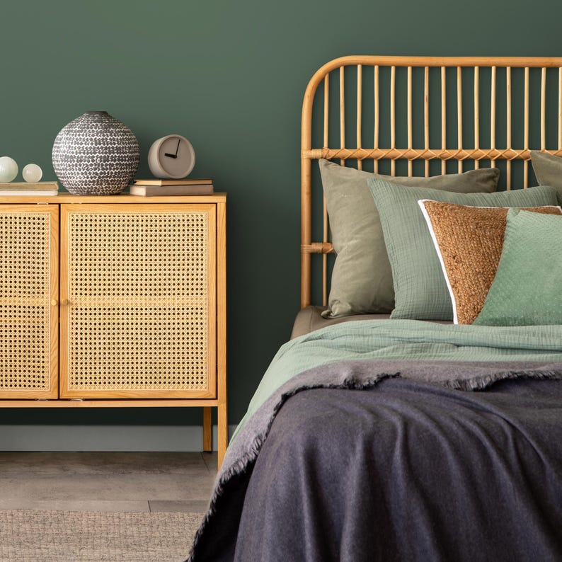 May include: A rattan headboard with a green and brown patterned throw blanket on a bed with a dark gray blanket. A wooden cabinet with a woven rattan door is in the foreground.