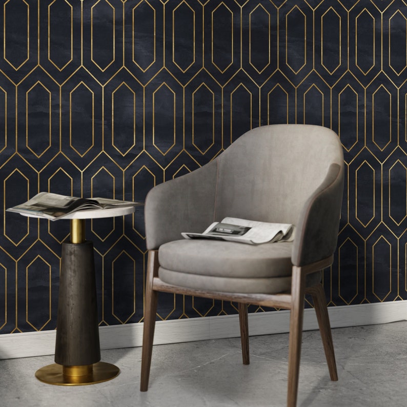 May include: A gray upholstered armchair with wooden legs sits in front of a dark blue wall with a gold geometric pattern. A small round table with a white top and a dark wood base sits to the left of the chair.