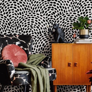 May include: A room scene featuring a black and white patterned wallpaper. A black velvet armchair with floral and bird designs is adorned with a round, dusty rose pillow and a green throw. A wooden cabinet holds a black monkey statue and a potted plant.