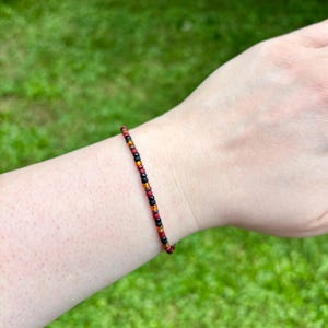 May include: A delicate beaded bracelet featuring a repeating pattern of small, round beads in matte black, deep red, and metallic copper tones. The beads are strung together in a simple, continuous line, creating a slim and understated accessory.