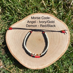 May include: Two beaded bracelets with adjustable black cords. One bracelet features ivory and gold beads, while the other has red and black beads. The text on the image reads "Morse Code: Angel - Ivory/Gold, Demon - Red/Black."