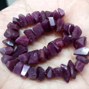 May include: A string of deep purple gemstone beads, each with an irregular, faceted shape. The beads, in varying shades of dark violet, are strung together. The gemstones are displayed, forming a circular shape.