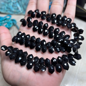 May include: A hand holds strands of glossy, faceted black beads, possibly gemstones. The beads are strung together. Other blue beads are visible in the background. The beads are likely measured in centimetres.