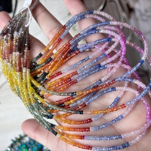 May include: A hand displays strands of multi-coloured gemstone beads. The beads are in shades of yellow, orange, red, blue, and purple. The beads are small and faceted, and appear to be strung together. The background is blurred.