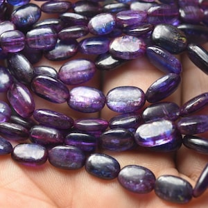May include: A strand of oval-shaped, iridescent purple gemstone beads. The beads have a shimmery, almost metallic finish.