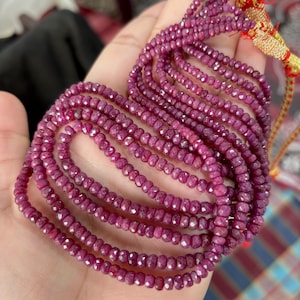 Op de afbeelding: Een verzameling robijn edelsteen kettingen. De kettingen hebben een dieprode kleur en zijn gemaakt van kleine, facetgeslepen kralen. De kettingen worden in een hand gehouden, met de uiteinden samengebonden met goud en rode draad.