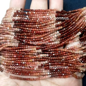 May include: A strand of red and pink gemstone beads, with a square shape, and a faceted finish. The beads are strung on a clear thread and are approximately 2mm in size. The beads are arranged in a loose pile, with some of the beads overlapping. The beads are a mix of red, pink, and clear colors.