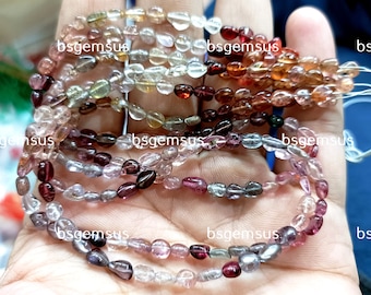 May include: A strand of multi-colored gemstone beads, with a variety of shades of pink, red, brown, and clear. The beads are oval shaped and have a smooth, polished finish. The strand is labeled "bsgemsus" in multiple places.