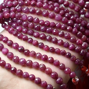 Op de afbeelding: Close-up van meerdere strengen gefacetteerde, dieprode robijn kralen. Elke kraal is rond en heeft een diameter van ongeveer 0,5 cm. De kralen zijn geregen, met rode kwastjes en zilverkleurige accenten aan de uiteinden.