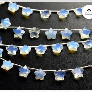 Könnte beinhalten: Eine Strähne aus weißen Opal-Sternenperlen auf einem silbernen Draht. Die Perlen sind facettiert und haben eine milchig weiße Farbe mit einem leichten irisierenden Schimmer.